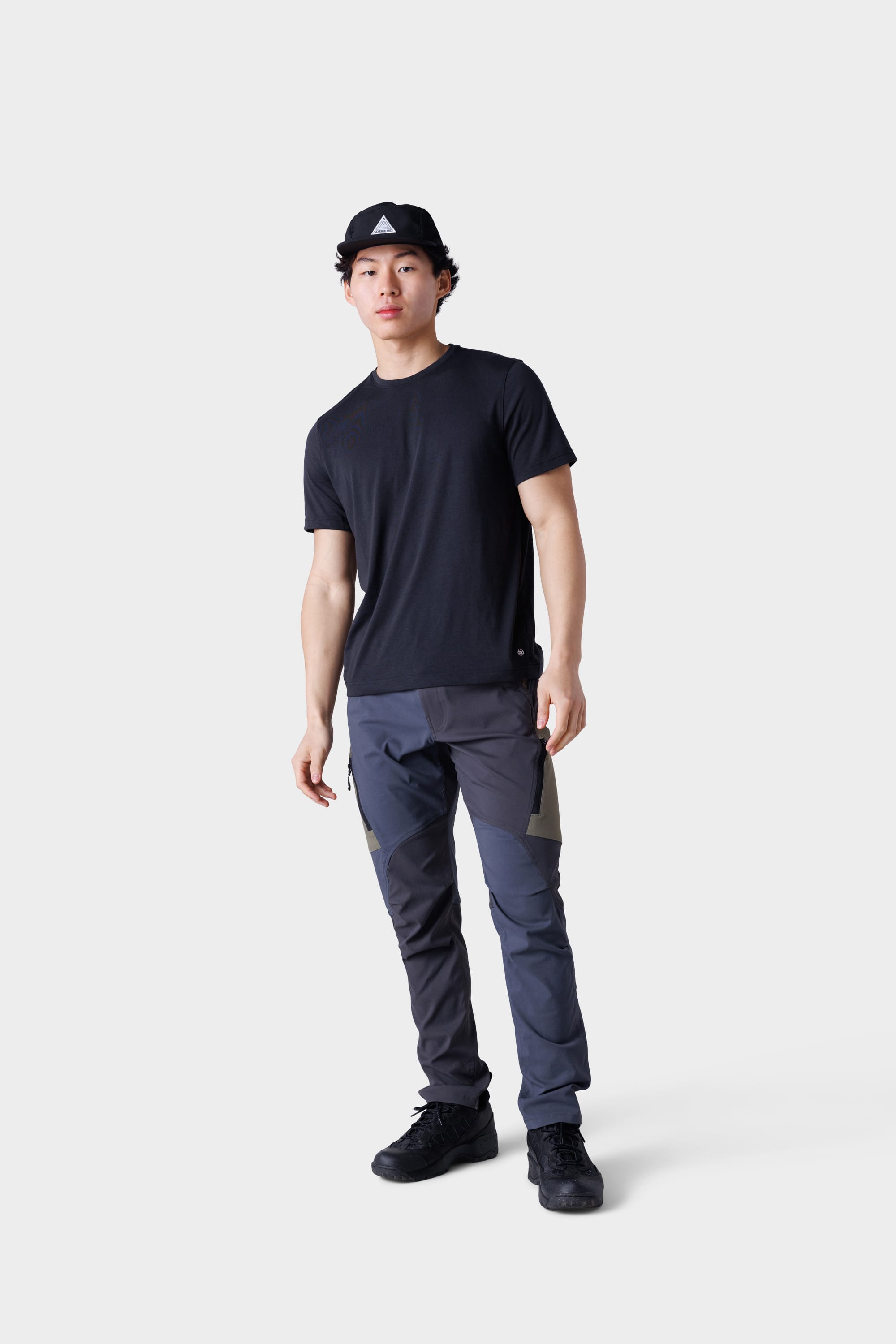 A young man in a black cap, black t-shirt, and 686 Men's Anything Cargo Pant - Slim Fit with a 13-pocket design stands against a white background, facing forward in black shoes and a relaxed pose, ready for any adventure.