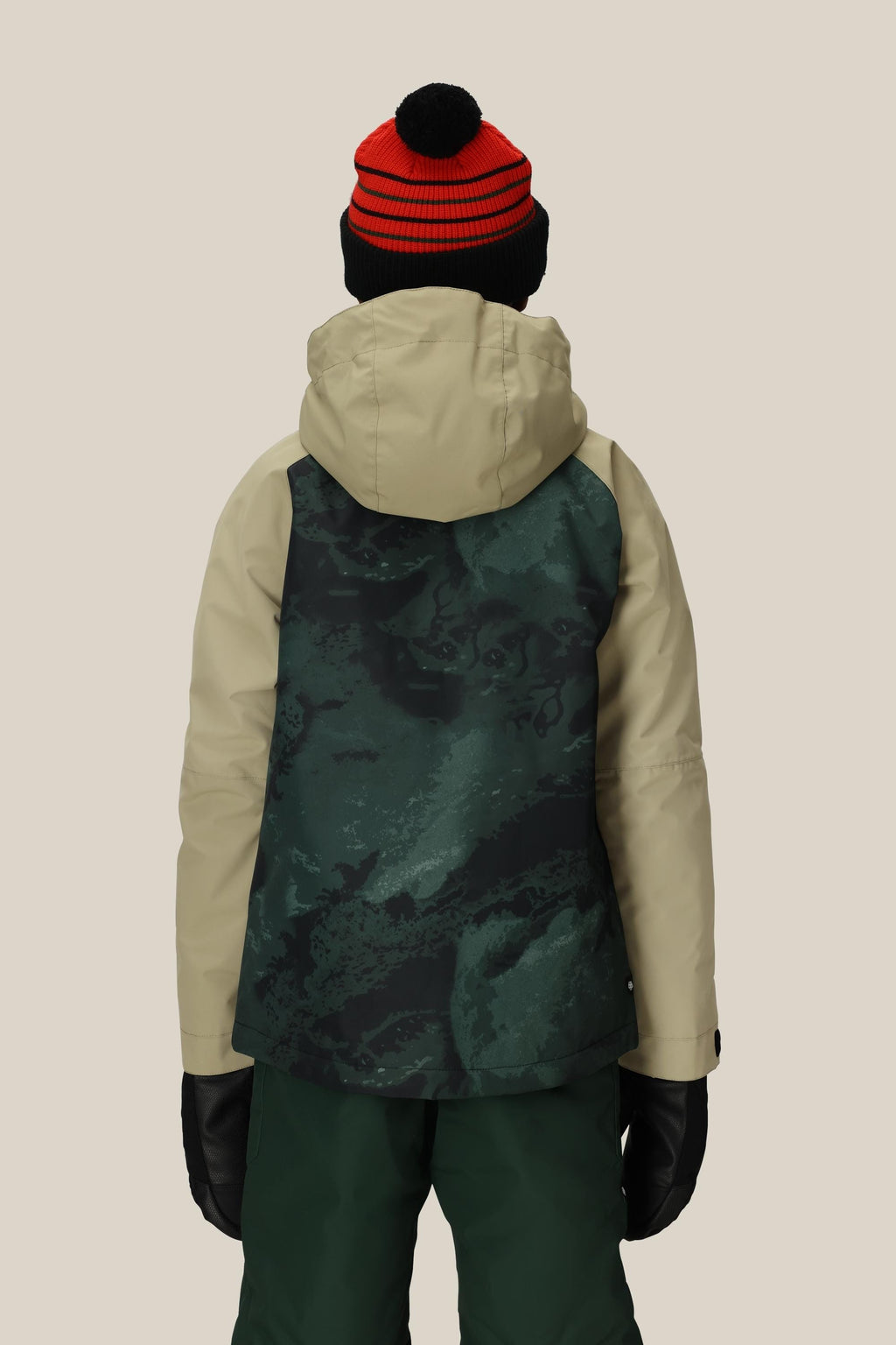A child wearing the 686 Boys' Hydra™ Insulated Jacket in beige and green, paired with green pants, black gloves, and a red and black striped beanie, stands facing away against a plain beige background.