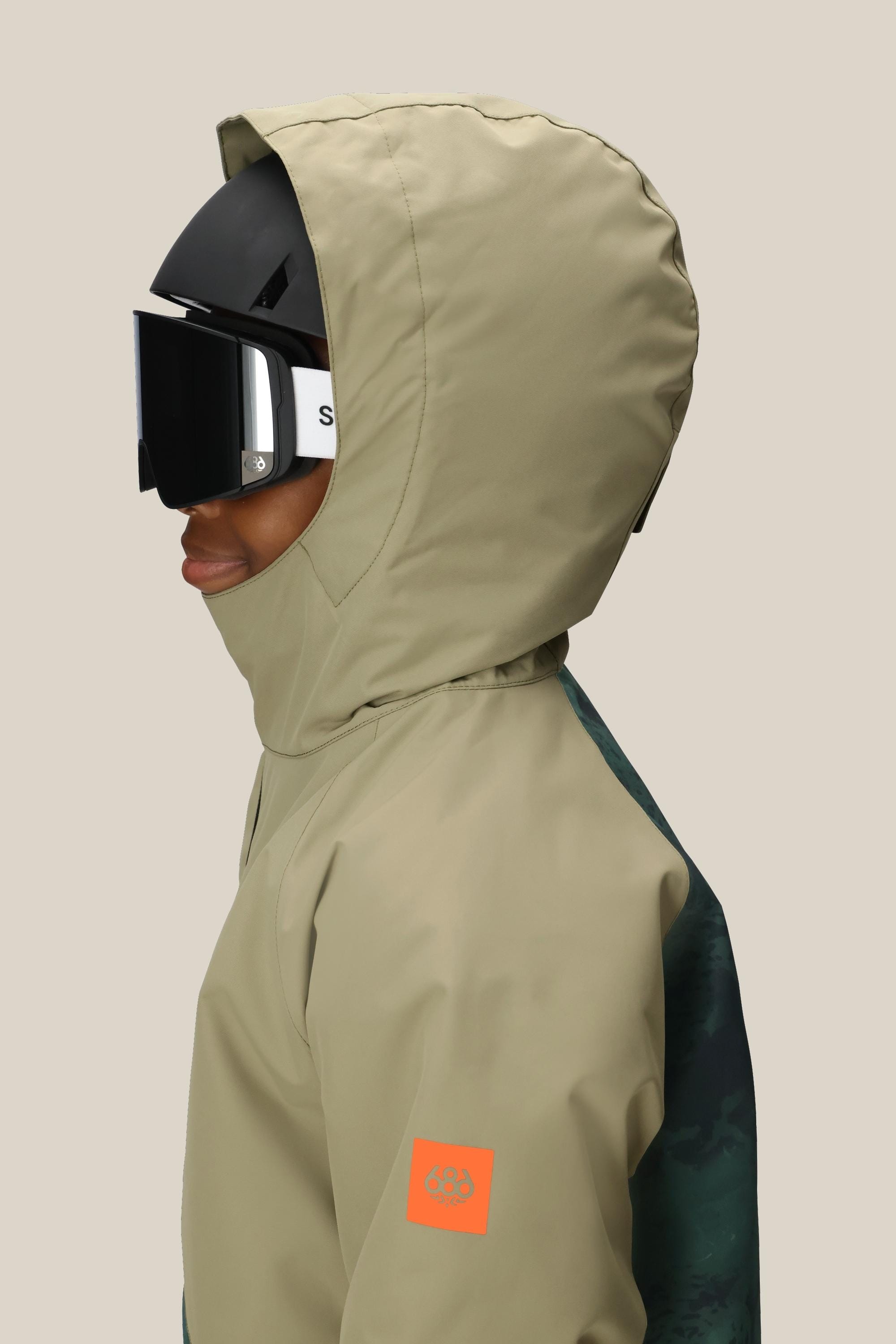 A boy in the 686 Boys' Hydra™ Insulated Jacket, tan with an orange patch and body-mapped lining, wears a black ski helmet and large black goggles, shown in profile against a plain light background. Brand: 686.
