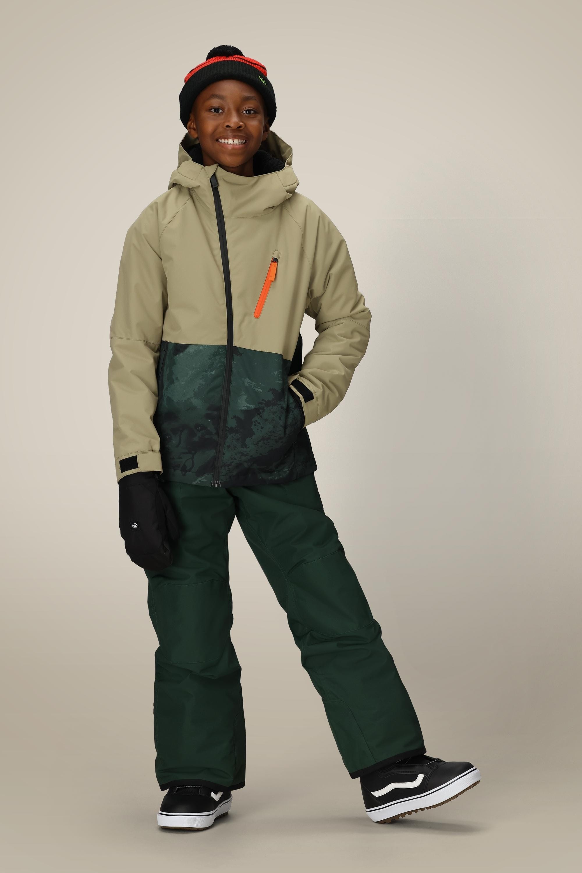 A smiling child in the 686 Boys' Hydra™ Insulated Jacket stands with one leg bent against a plain background, also wearing snow pants, a beanie hat, gloves, and black-and-white shoes.