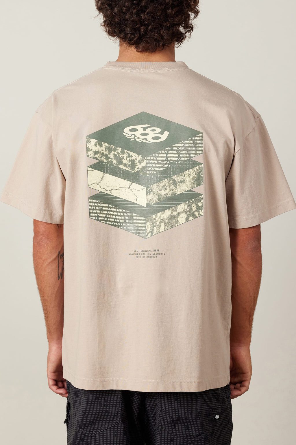 A person wears the 686 Elements Relaxed S/S Tee by 686, a beige men’s t-shirt made from 100% cotton, featuring geometric hexagonal graphics and layered abstract patterns with a small text section on the back.