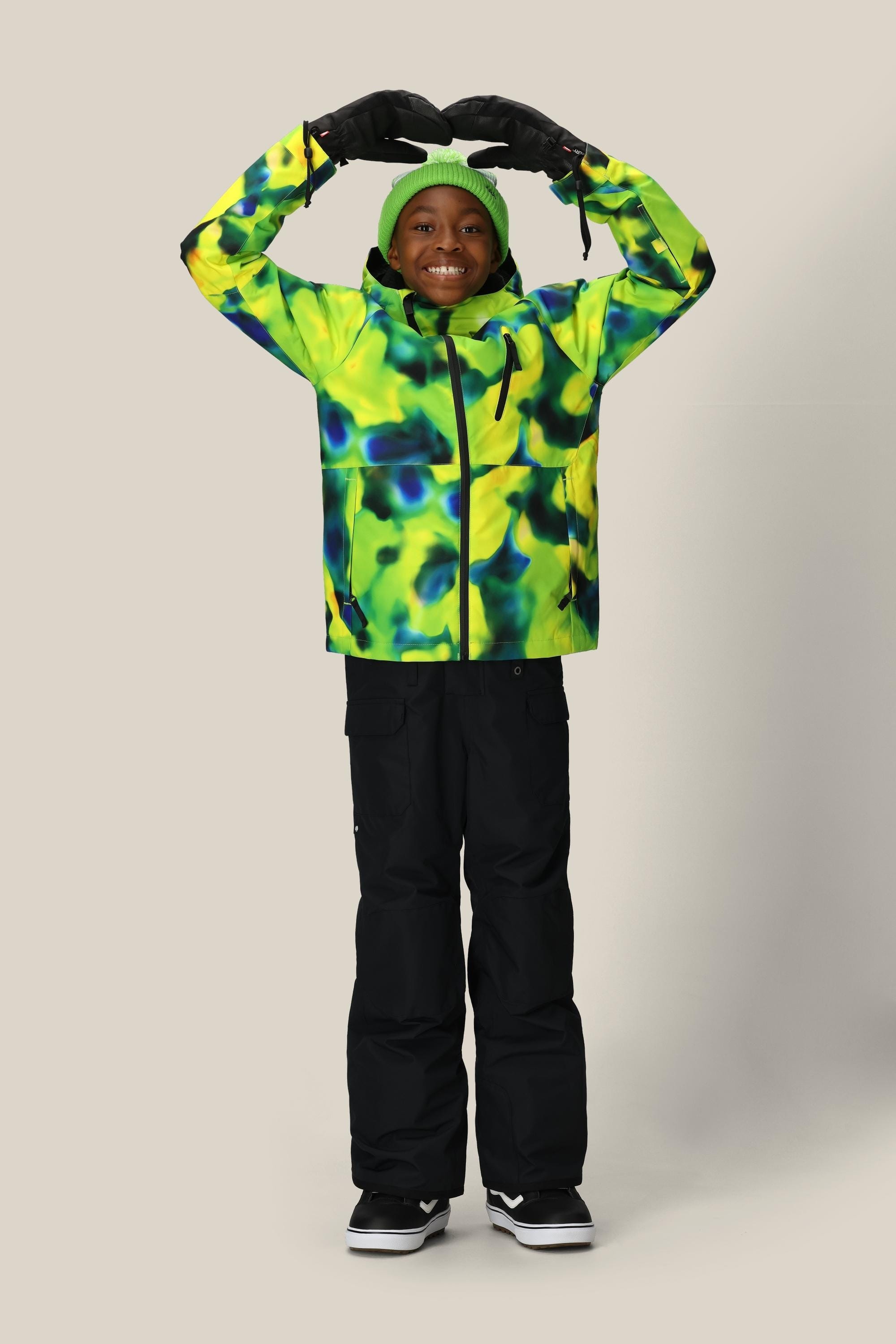 A smiling child stands against a plain background, arms raised in a heart shape, wearing the 686 Boys' Frontier™ Insulated Bib by 686, along with a bright yellow and green jacket, black gloves, and a green beanie.