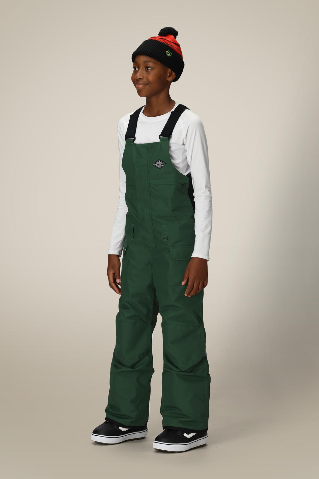 A smiling child wears the 686 Boys' Frontier™ Insulated Bib in dark green, paired with a white long-sleeve shirt, a black-and-red beanie, and black sneakers with white soles, standing against a light plain background.
