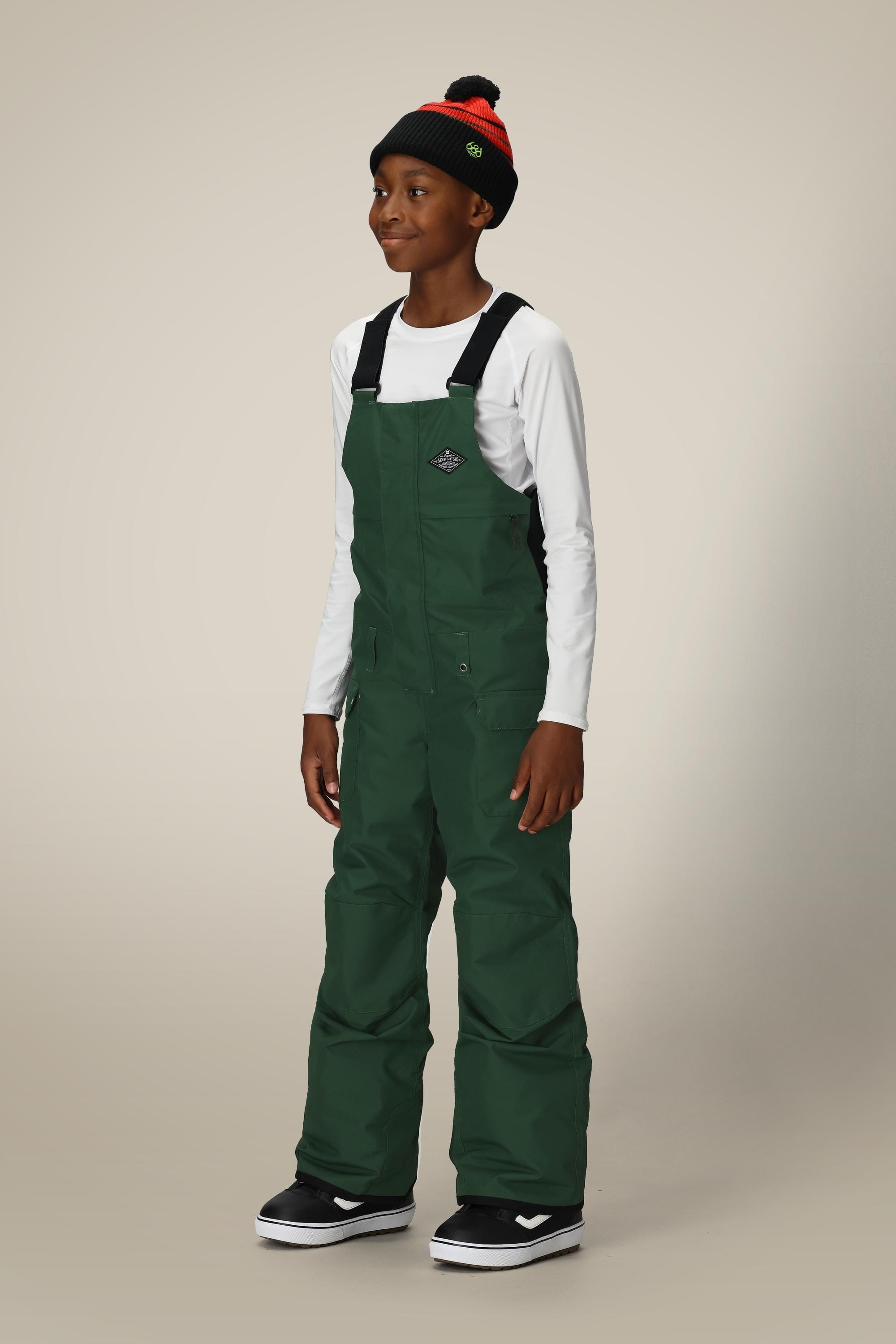 A smiling child wears the 686 Boys' Frontier™ Insulated Bib in dark green, paired with a white long-sleeve shirt, a black-and-red beanie, and black sneakers with white soles, standing against a light plain background.