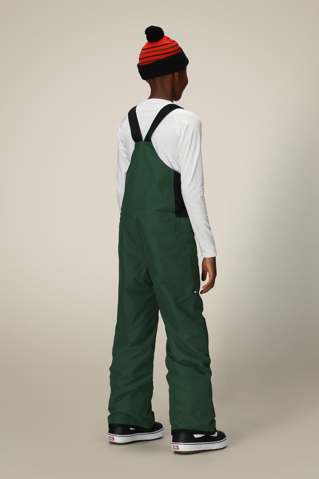 A child stands facing away against a plain light background, wearing a red and black striped beanie, a white long-sleeve shirt, and green 686 Boys' Frontier™ Insulated Bib overalls with extra pockets, paired with black sneakers with white soles.