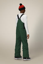 A child stands facing away against a plain light background, wearing a red and black striped beanie, a white long-sleeve shirt, and green 686 Boys' Frontier™ Insulated Bib overalls with extra pockets, paired with black sneakers with white soles.