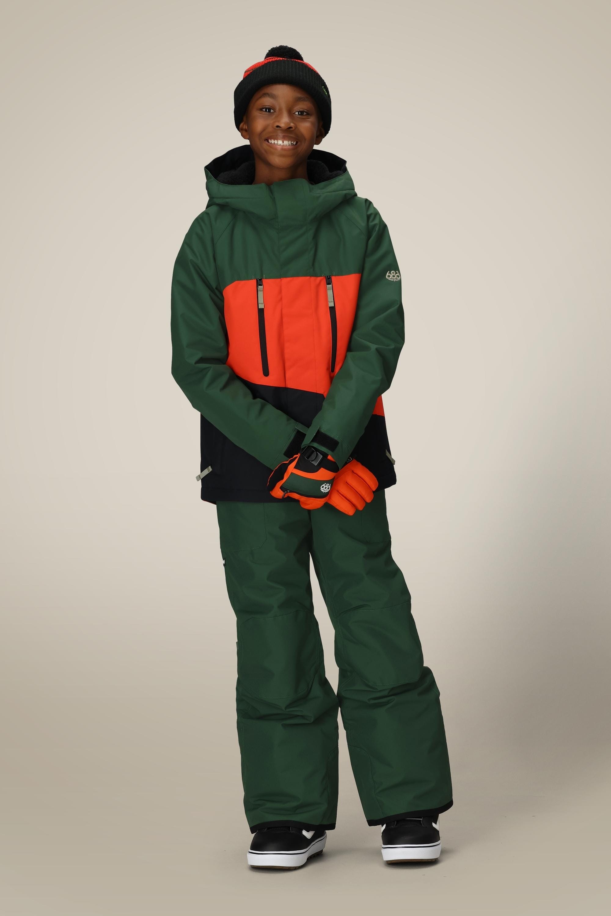 A smiling child stands wearing the 686 Boys' Frontier™ Insulated Bib, paired with a green and orange winter jacket, orange gloves, a black beanie with red stripe, and black snow boots against a plain background.