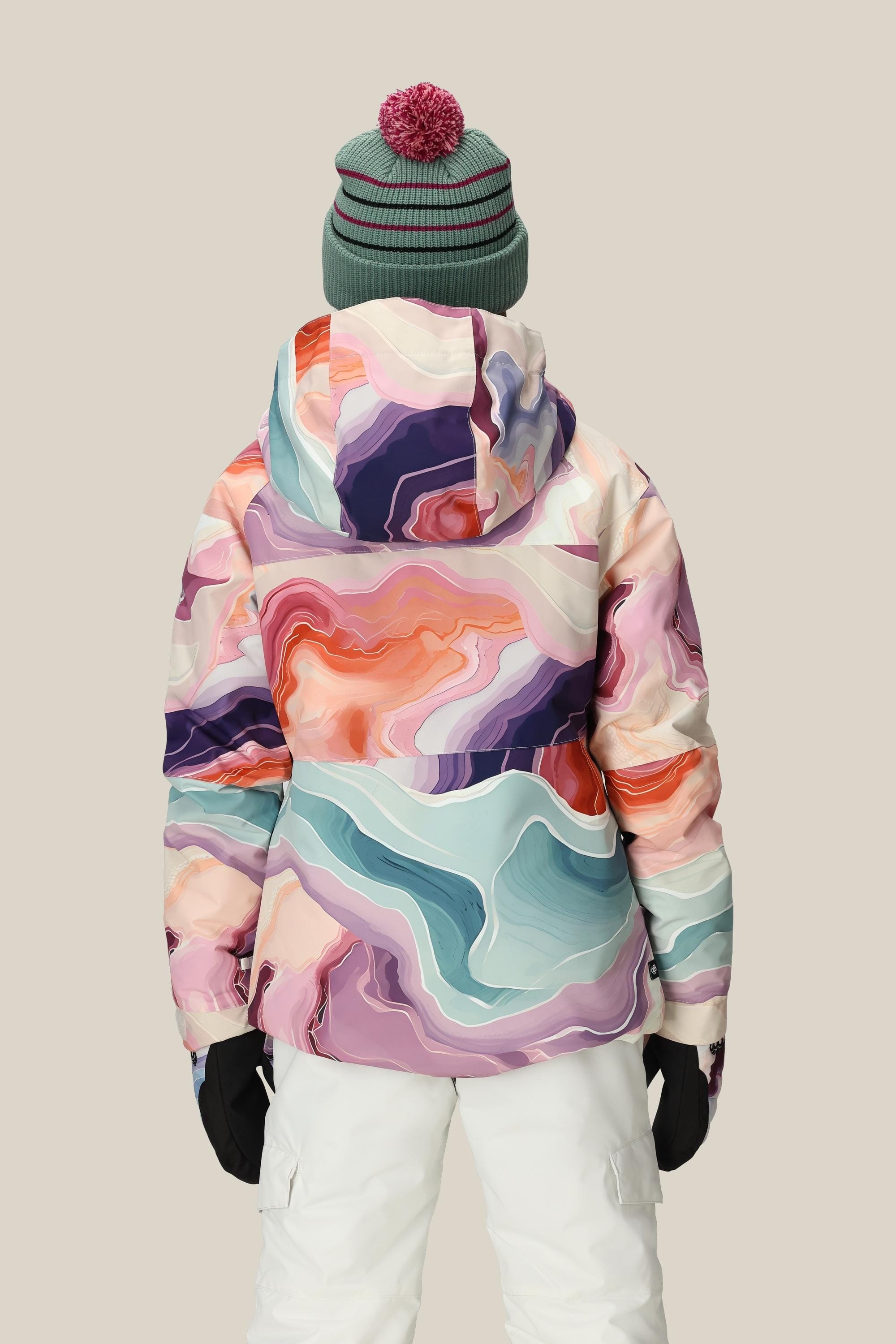 Someone wearing the 686 Girls' Athena™ Insulated Jacket stands with their back to the camera, paired with white snow pants, black gloves, and a green beanie featuring a red pom-pom against a plain background.