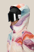 A person in the 686 Girls' Athena™ Insulated Jacket, featuring a colorful abstract pattern and hood, with a black ski helmet and large black goggles, shown from the side against a plain light beige background.