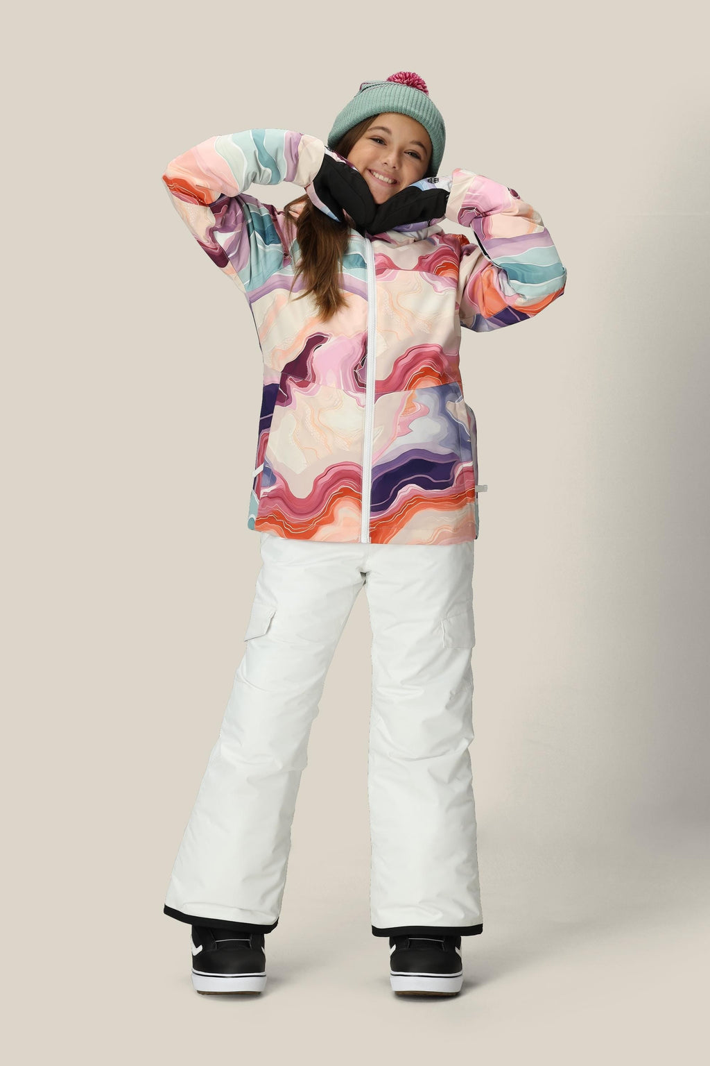 A person smiles with hands raised, wearing the 686 Girls' Athena™ Insulated Jacket by 686, a green beanie, white snow pants, and black gloves against a plain beige background.