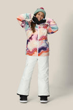 A person smiles with hands raised, wearing the 686 Girls' Athena™ Insulated Jacket by 686, a green beanie, white snow pants, and black gloves against a plain beige background.