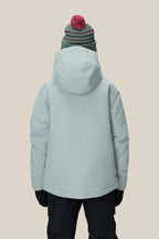 A girl wears the 686 Girls' Hydra™ Insulated Jacket in light blue, paired with dark gloves, navy pants, and a green beanie with a red pom-pom, standing against a beige background and prepared for chilly weather.