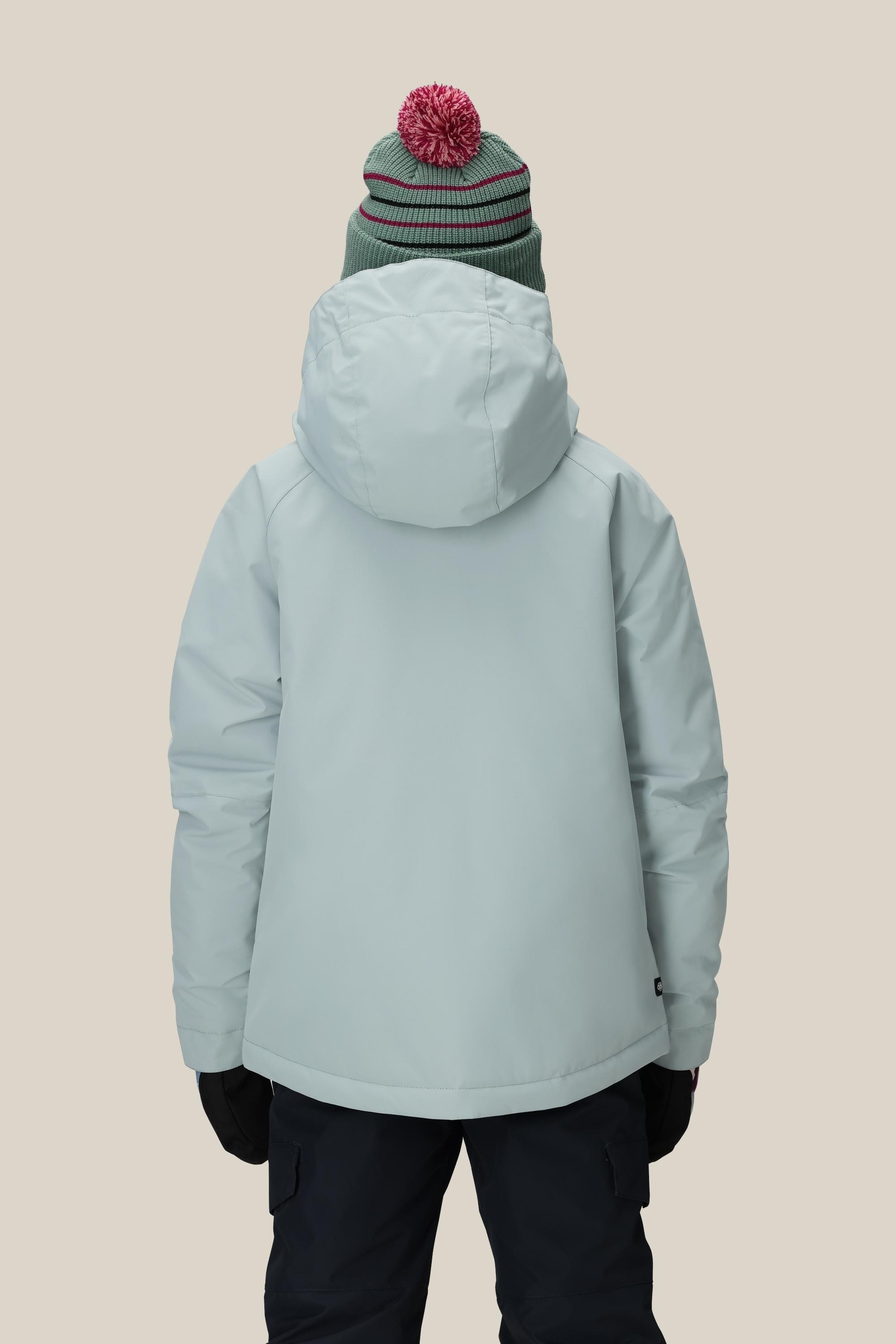 A girl wears the 686 Girls' Hydra™ Insulated Jacket in light blue, paired with dark gloves, navy pants, and a green beanie with a red pom-pom, standing against a beige background and prepared for chilly weather.