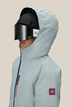 A girl wearing the 686 Girls' Hydra™ Insulated Jacket in light blue with body-mapped lining, chest zipper, and a small sleeve patch, paired with a black ski helmet and goggles. Side view against a beige background.