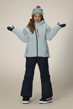 A young girl stands smiling with arms out against a plain background, wearing the 686 Girls' Hydra™ Insulated Jacket in light blue, dark snow pants, gloves, and a pom-pom knit hat—radiating warmth in her 686 outerwear.