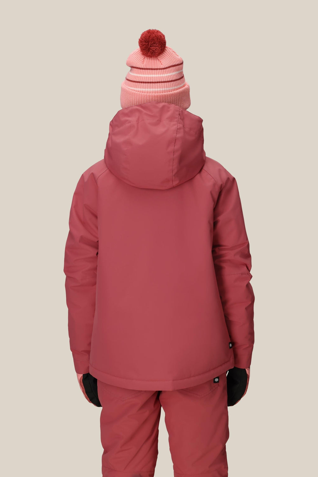 Wearing the 686 Girls' Hydra™ Insulated Jacket by 686, matching pants, black gloves, and a pink beanie with a red pompom, a person stands facing away from the camera against a plain beige background.