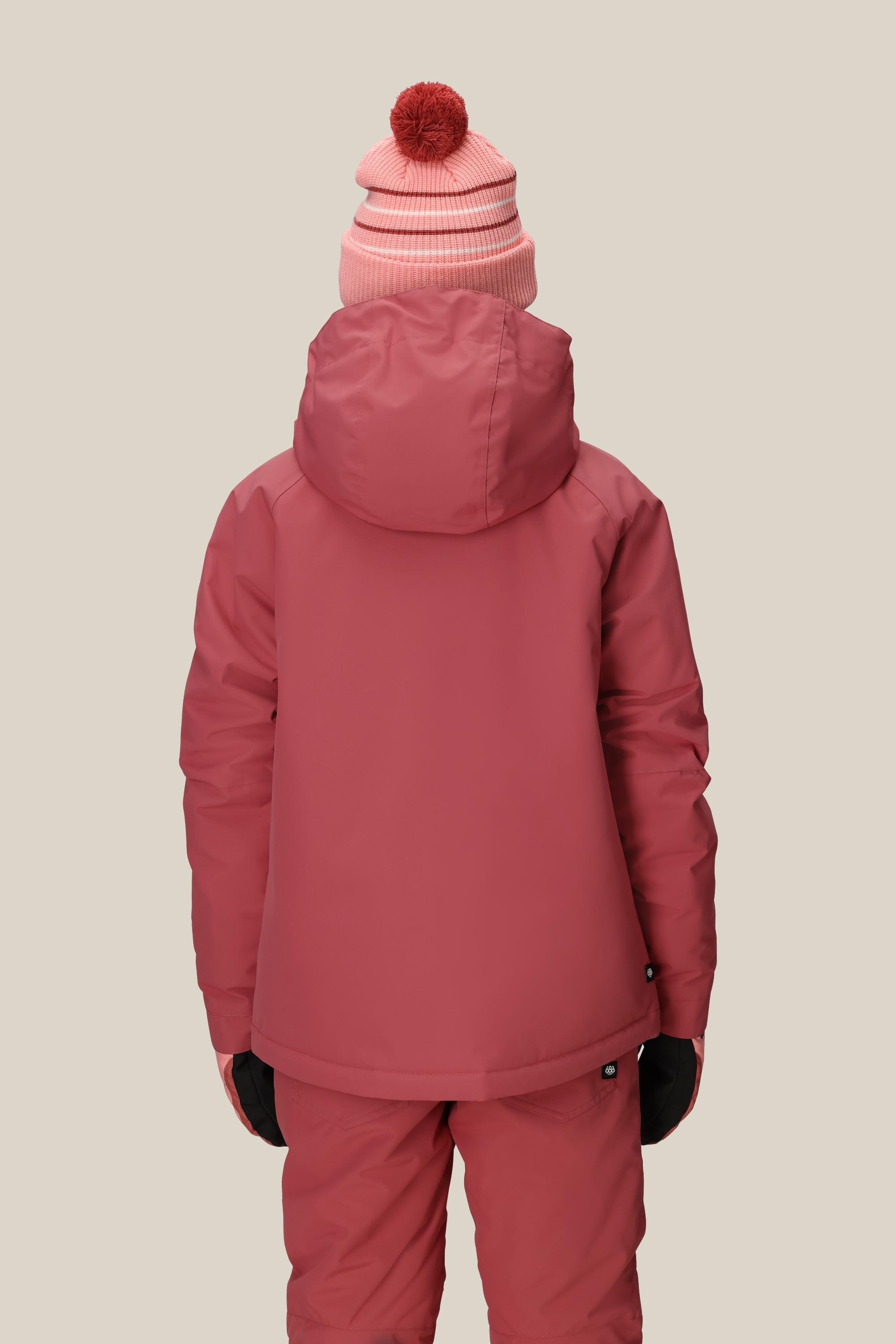 Wearing the 686 Girls' Hydra™ Insulated Jacket by 686, matching pants, black gloves, and a pink beanie with a red pompom, a person stands facing away from the camera against a plain beige background.