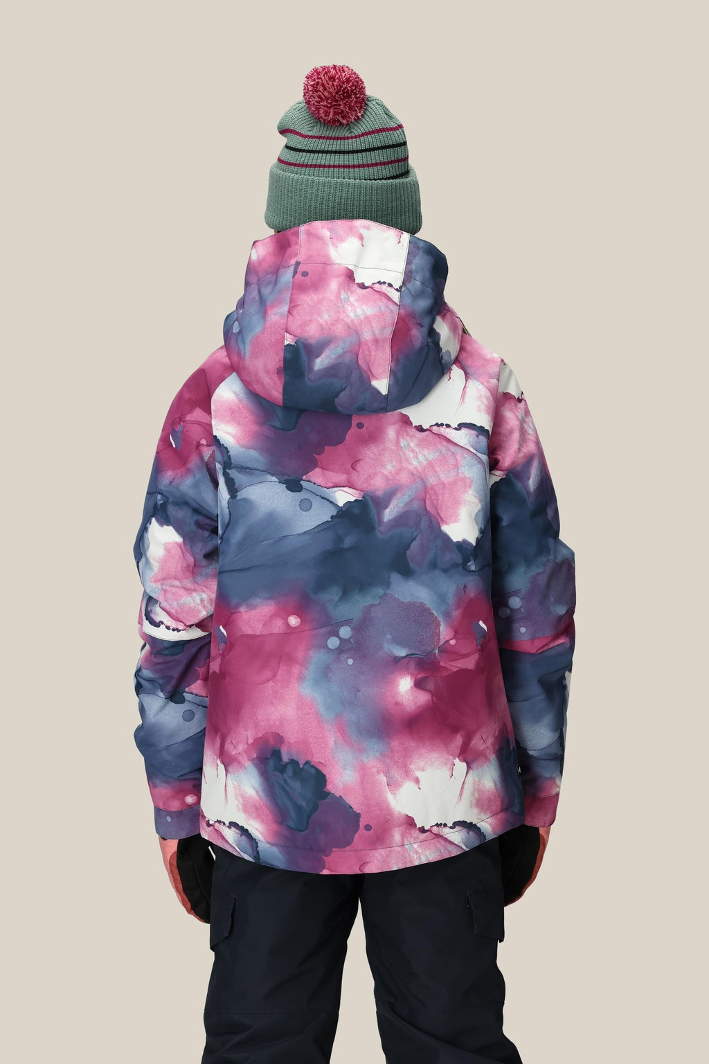 A person is seen from behind wearing the 686 Girls' Hydra™ Insulated Jacket by 686 in a blue, pink, and white watercolor pattern with body-mapped lining for warmth, paired with black pants, black gloves, and a green knit beanie with a red pom-pom.