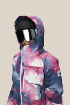 A girl smiles in the 686 Girls' Hydra™ Insulated Jacket—featuring a vibrant, watercolor pattern and hood—paired with a black ski helmet and reflective goggles, ready for warmth on any adventure against a beige background.