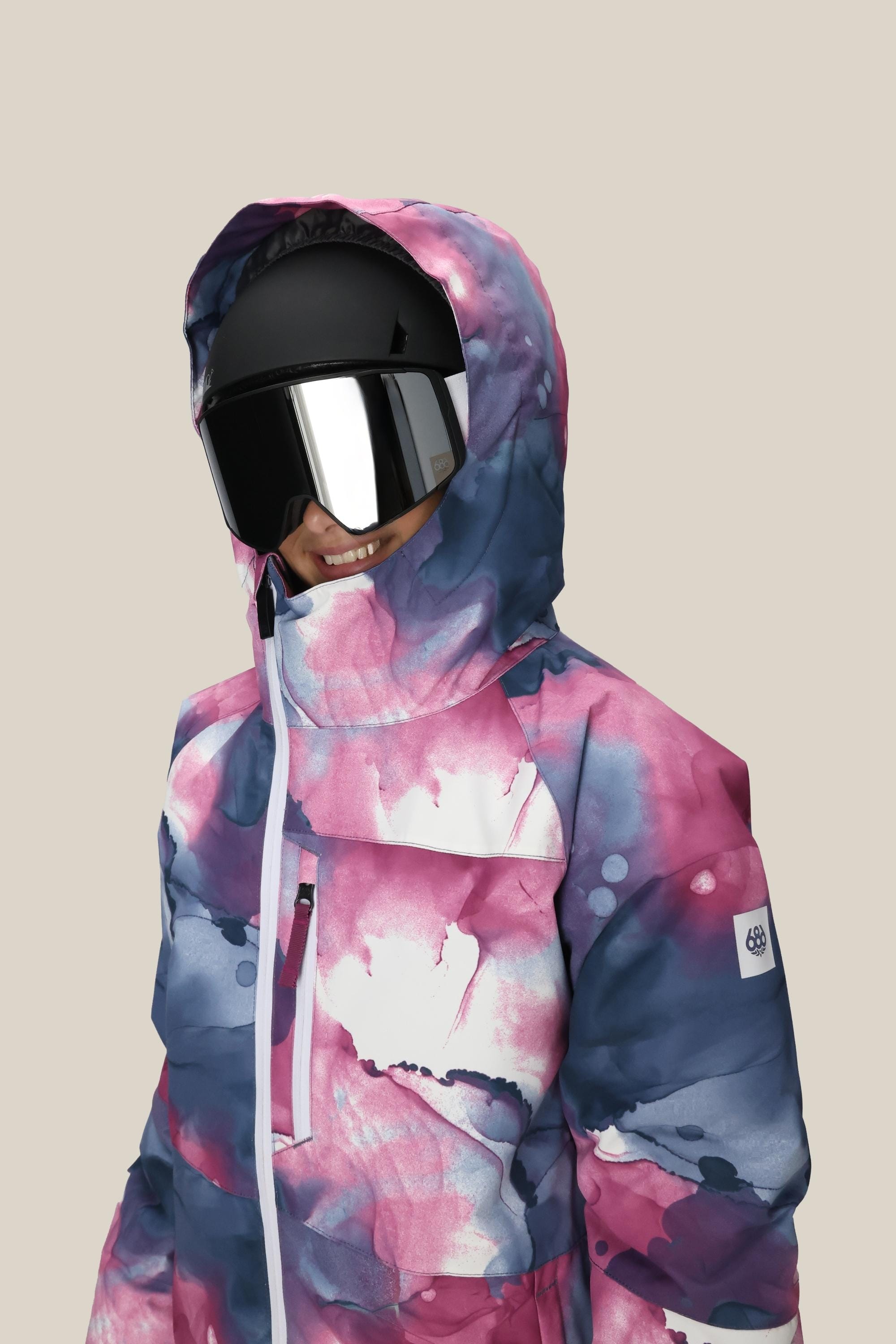 A girl smiles in the 686 Girls' Hydra™ Insulated Jacket—featuring a vibrant, watercolor pattern and hood—paired with a black ski helmet and reflective goggles, ready for warmth on any adventure against a beige background.