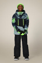 A smiling child wearing 686 Boys Infinity™ Cargo Insulated Pant, a green beanie, black and green winter jacket with neon accents, gloves, and black sneakers stands against a plain beige background.