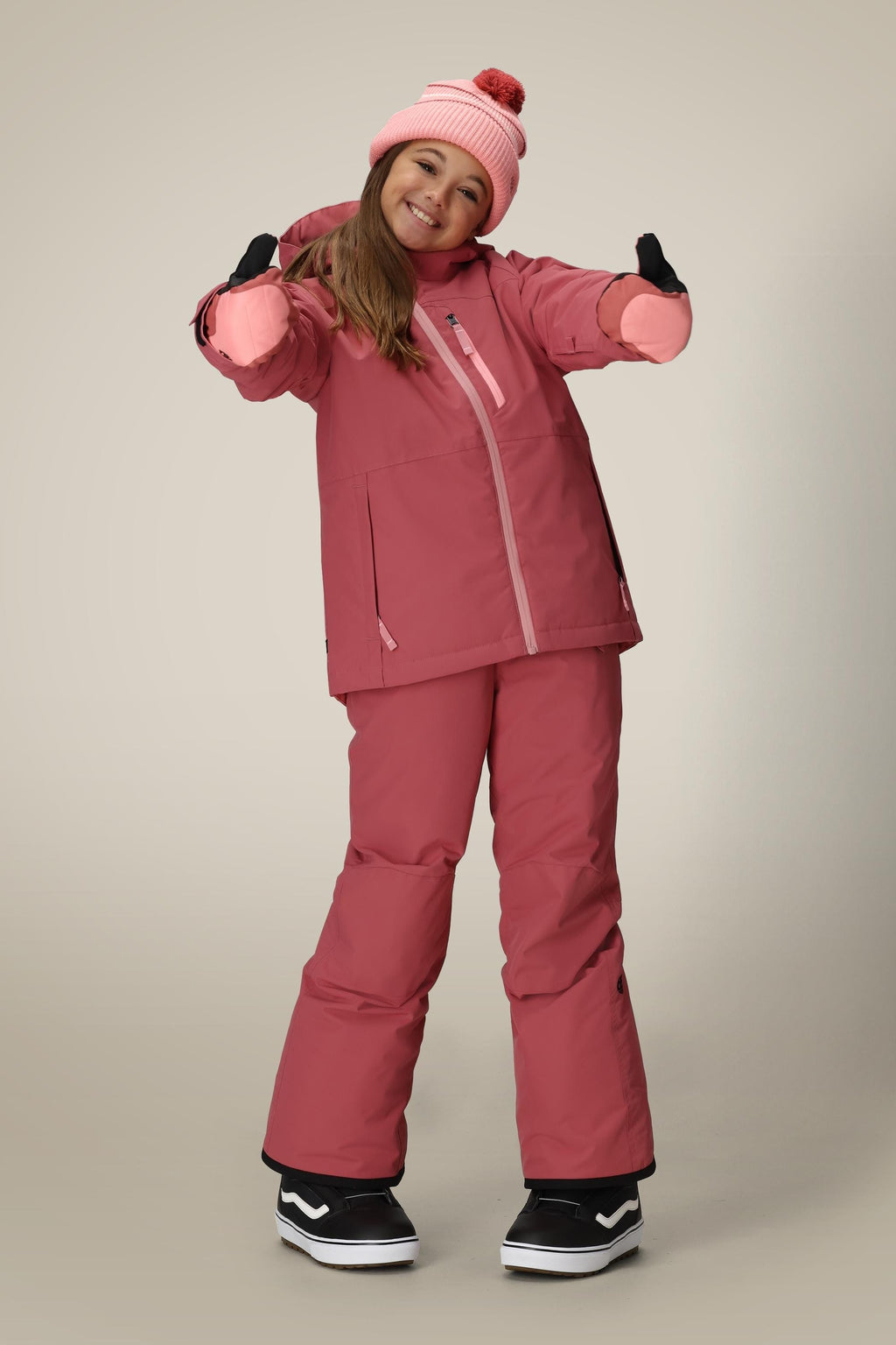 A smiling child stands with arms outstretched, wearing the 686 Girls Sierra Insulated Bib by 686, a pink jacket and beanie with pom-pom, matching pants, black gloves, and black-and-white boots against a neutral background.
