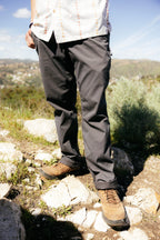 A person in 686 Men's Everywhere® Pant - Relaxed Fit stands on rocky ground outdoors with hills and greenery in the background under a blue sky, wearing brown hiking shoes and a light patterned shirt.