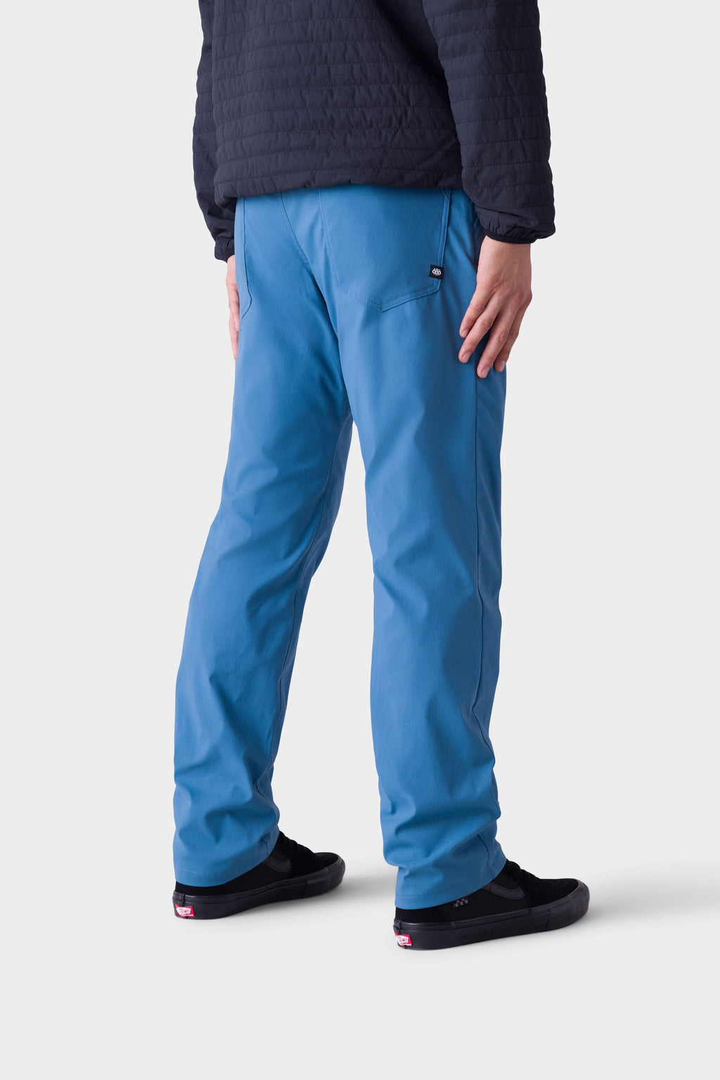 A person in a black quilted jacket, 686 Men's Everywhere® Merino-Lined Pant - Relaxed Fit by 686 in bright blue, and black slip-on shoes stands with their back to the camera against a plain white background.
