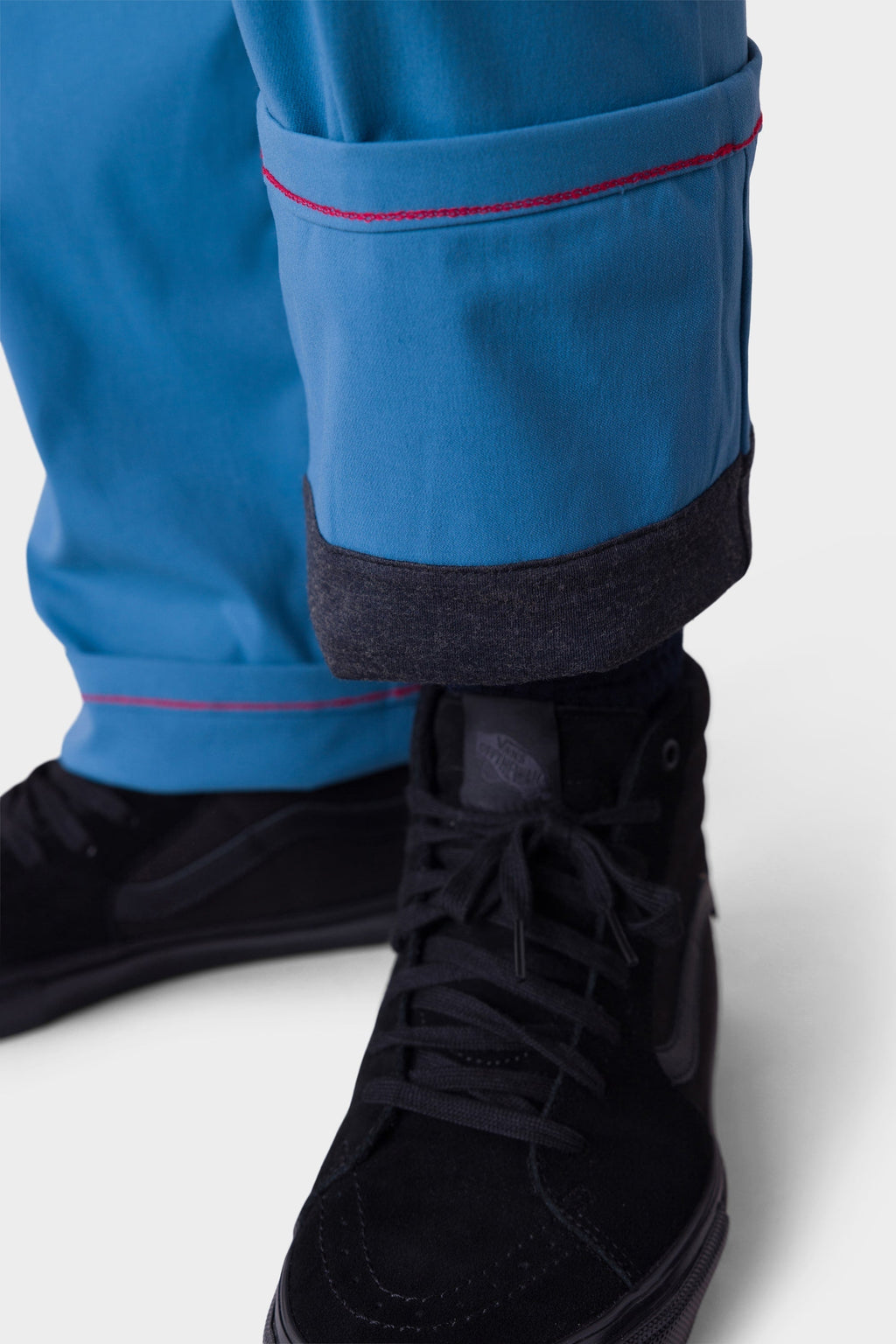 A close-up of a person wearing 686 Men's Everywhere® Merino-Lined Pant - Relaxed Fit in blue with red stitching, rolled cuffs revealing a dark merino lining, paired with black high-top lace sneakers and a stylish insulated outerwear piece for cold days.