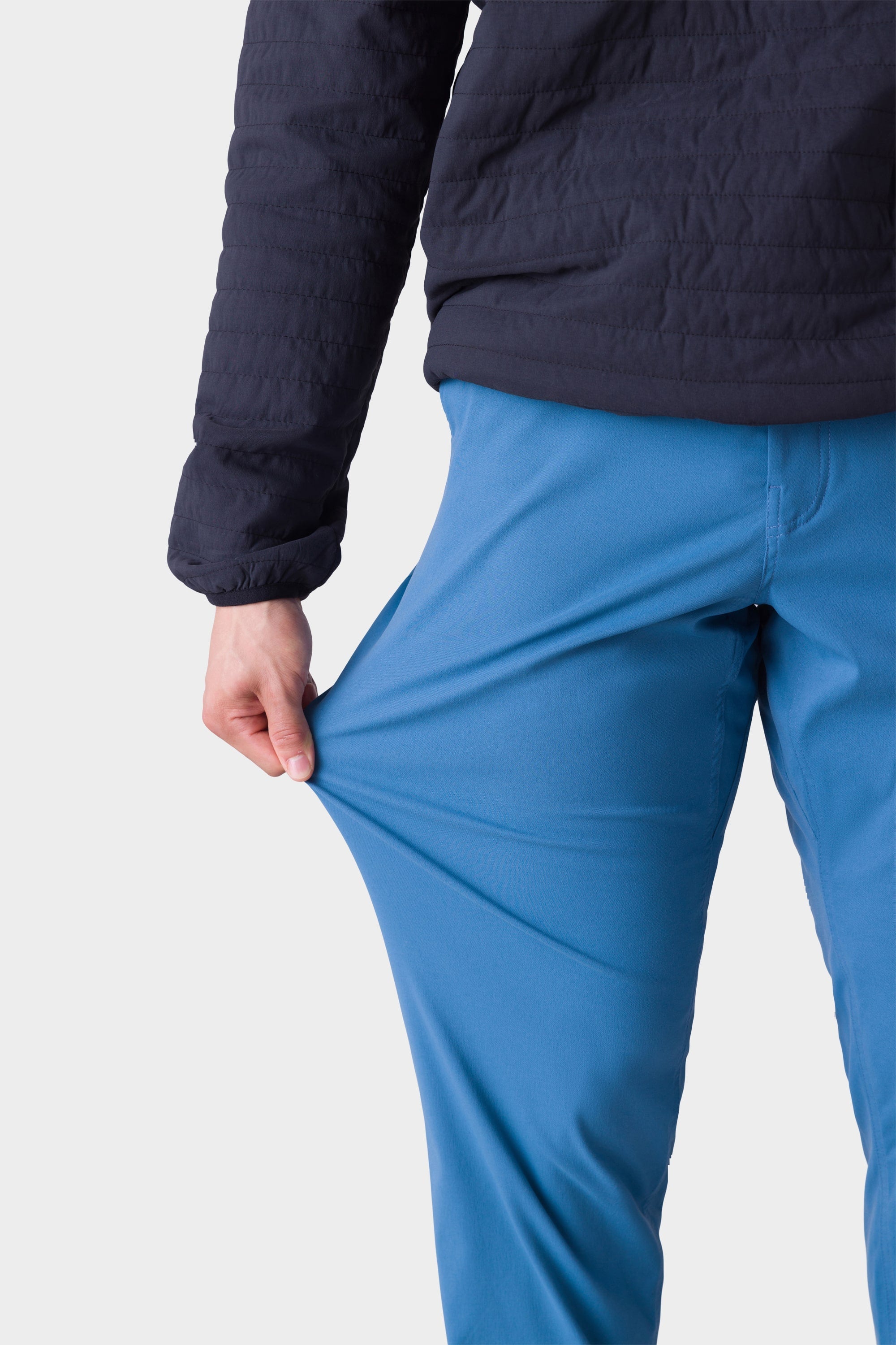 A person wearing a stylish insulated jacket and 686 Men's Everywhere® Merino-Lined Pant - Relaxed Fit stretches the pants' bright blue fabric to demonstrate its flexibility; the image focuses on their torso and legs.