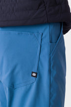 Close-up of a person wearing 686 Men's Everywhere® Merino-Lined Pant - Relaxed Fit, featuring a back pocket with a black and white logo tag. They pair it with an insulated jacket. Plain, light-colored background. Brand: 686.