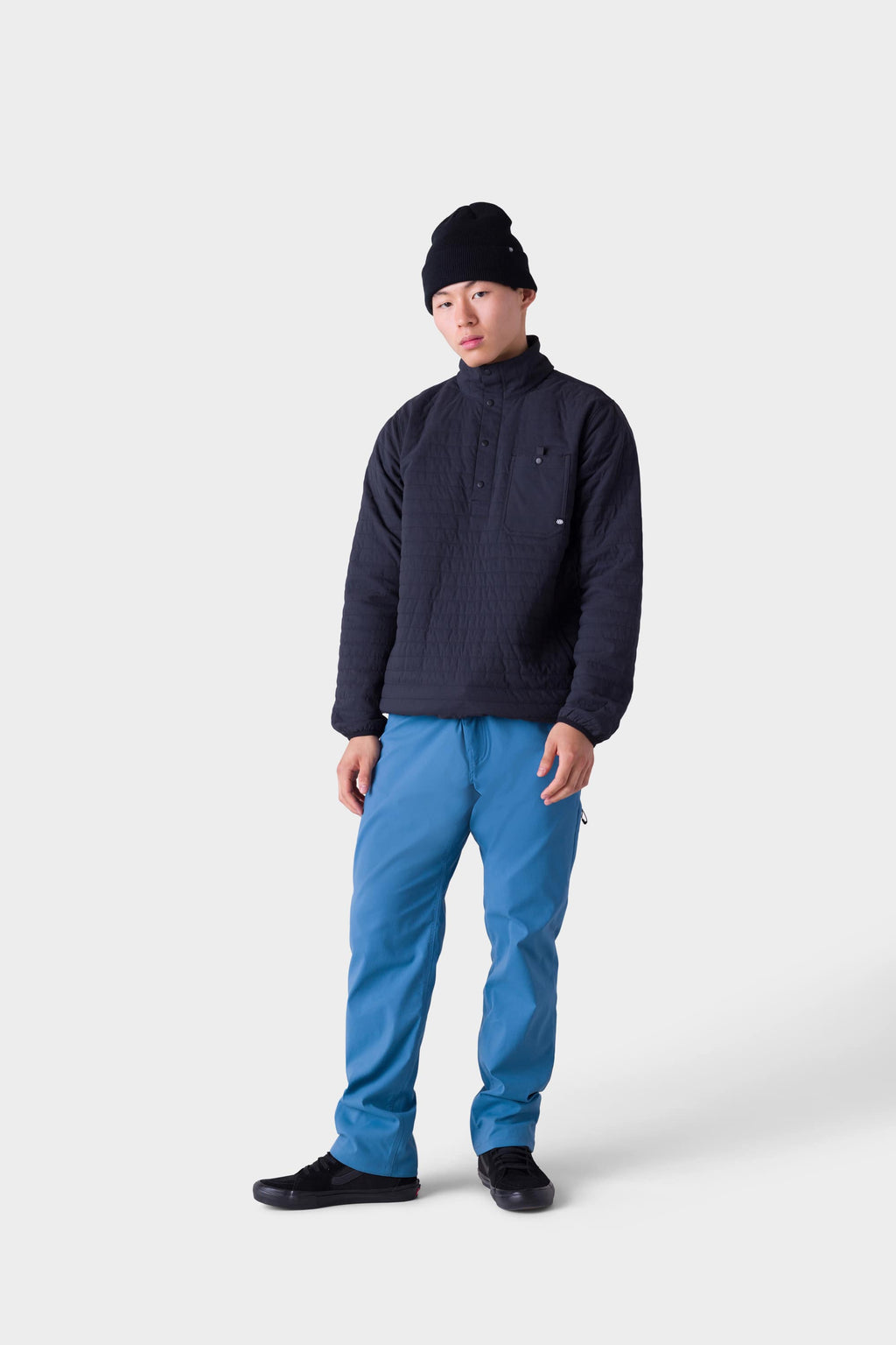 A person wearing a black beanie, stylish insulated outerwear, 686 Men's Everywhere® Merino-Lined Pant - Relaxed Fit in blue, and black shoes stands against a plain white background, looking at the camera with a neutral expression.