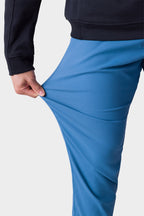 A man wearing the 686 Men's Everywhere® Merino-Lined Pant - Slim Fit stretches the blue pants' fabric with his left hand, highlighting their flexibility and warmth—ideal for cold weather.