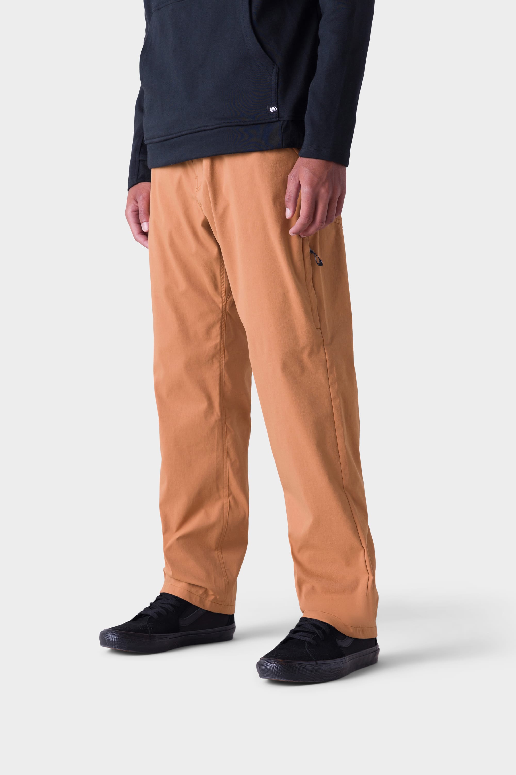 A person stands against a plain white background wearing 686 Men's Everywhere® Merino-Lined Pant - Relaxed Fit in light brown, a black hoodie under an insulated outerwear, and black sneakers. The image is cropped at the shoulders.