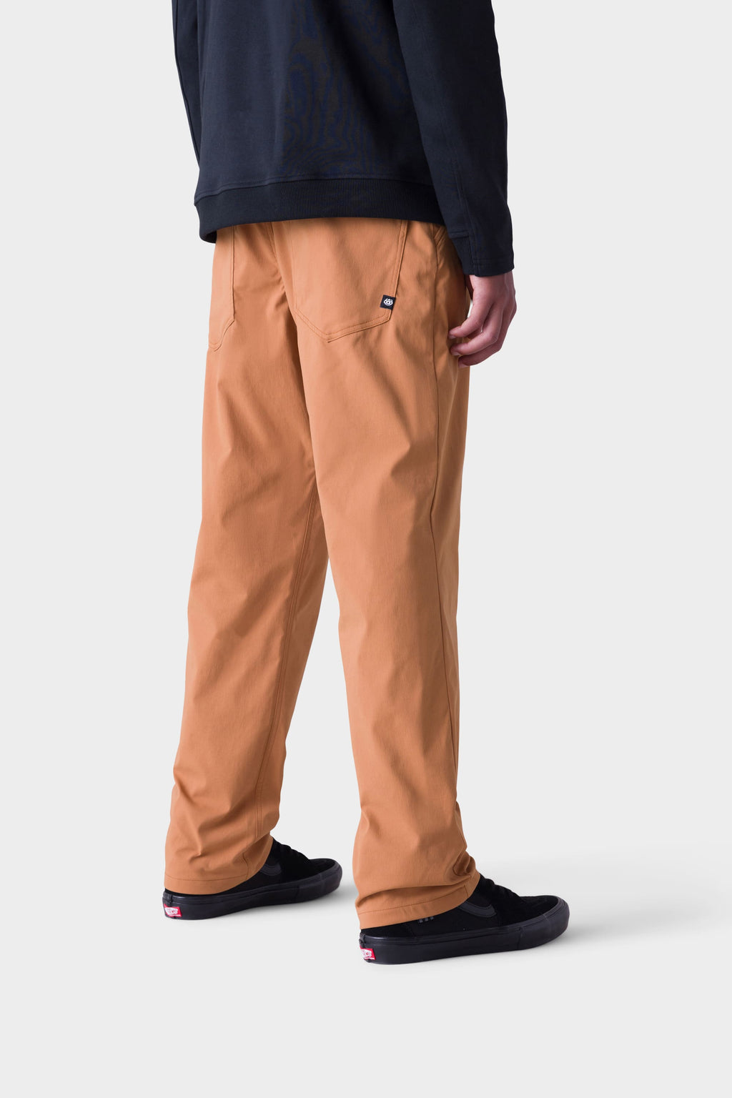 A person faces away against a plain light background, wearing 686 Men's Everywhere® Merino-Lined Pant - Relaxed Fit in tan, a black long-sleeve shirt, black sneakers, and stylish insulated outerwear.