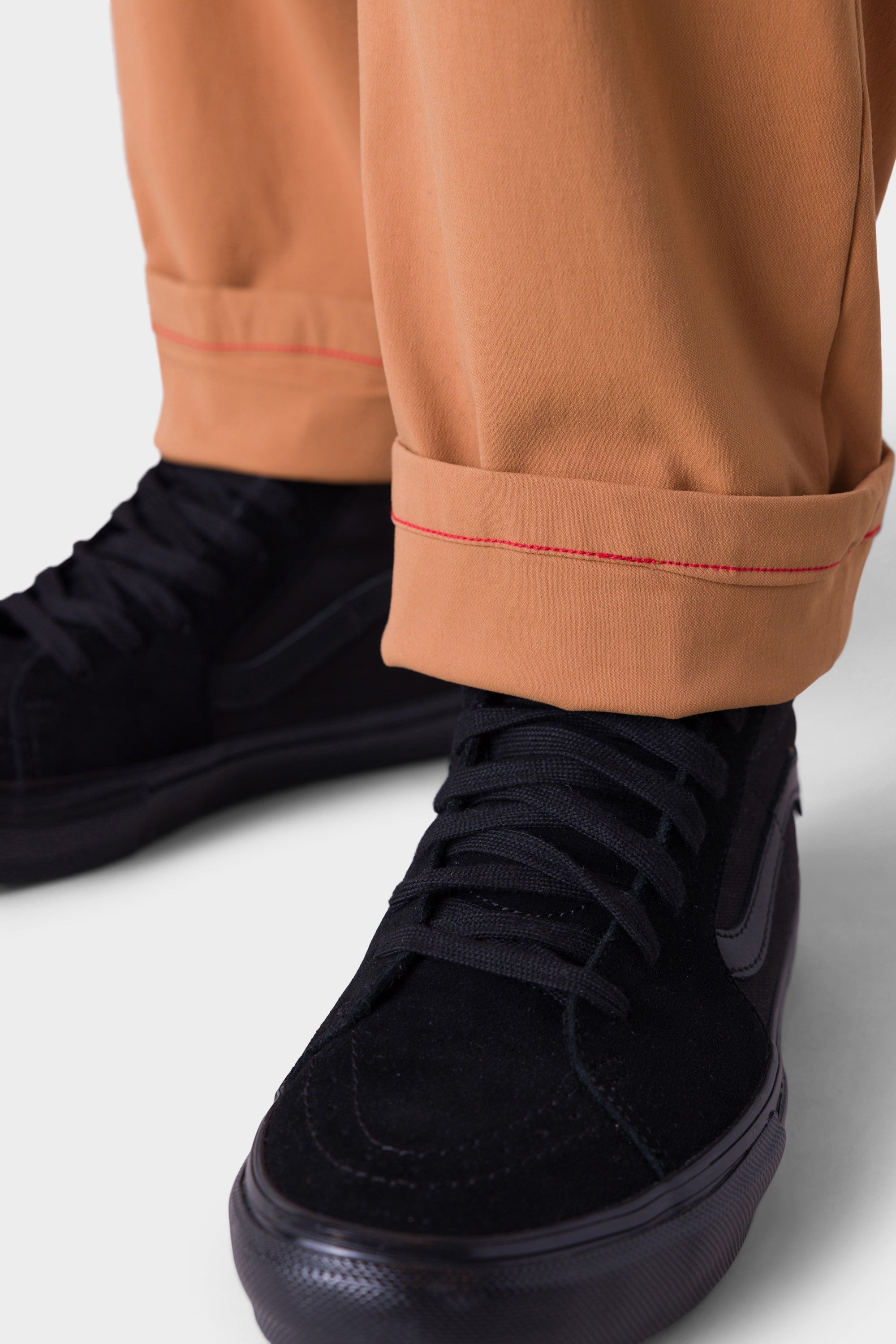 Close-up of a person wearing 686 Men's Everywhere® Merino-Lined Pant - Relaxed Fit in light brown, cuffed to reveal red stitching, paired with black suede sneakers—stylish and insulated for cold weather by 686.