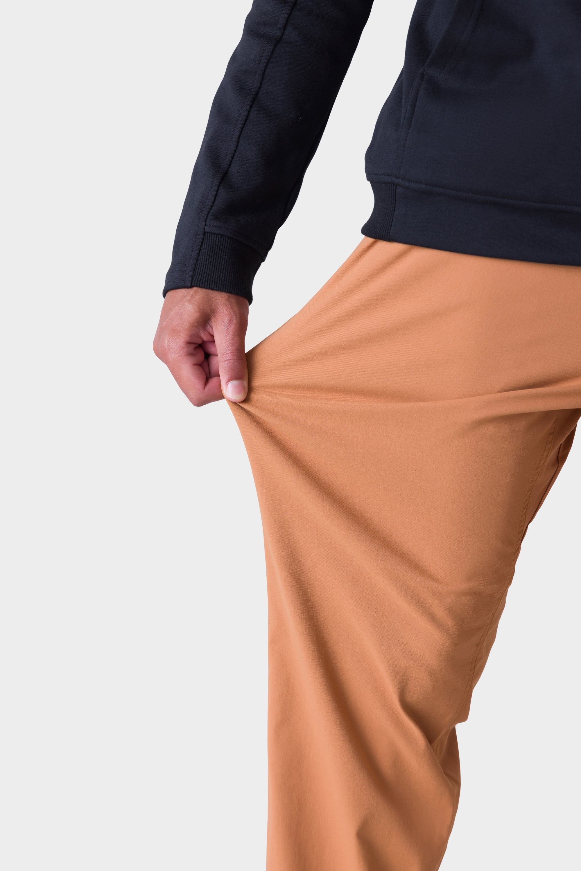 A person in a black long-sleeve top, tan 686 Men's Everywhere® Merino-Lined Pant - Relaxed Fit, and stylish insulated outerwear stretches the pants' fabric with one hand, showcasing the flexibility of this 686 product.