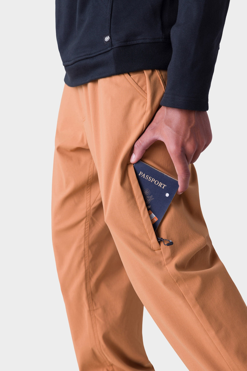 Wearing the 686 Men's Everywhere® Merino-Lined Pant - Relaxed Fit in tan and a dark top, a person slips a passport into the pants’ zippered side pocket. The background is plain and light-colored.