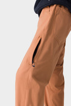 Close-up of a person wearing the 686 Men's Everywhere® Merino-Lined Pant - Relaxed Fit in light brown, featuring a partially open zippered thigh pocket. Only their lower body is visible, paired with a dark top and insulated outerwear against a plain background.