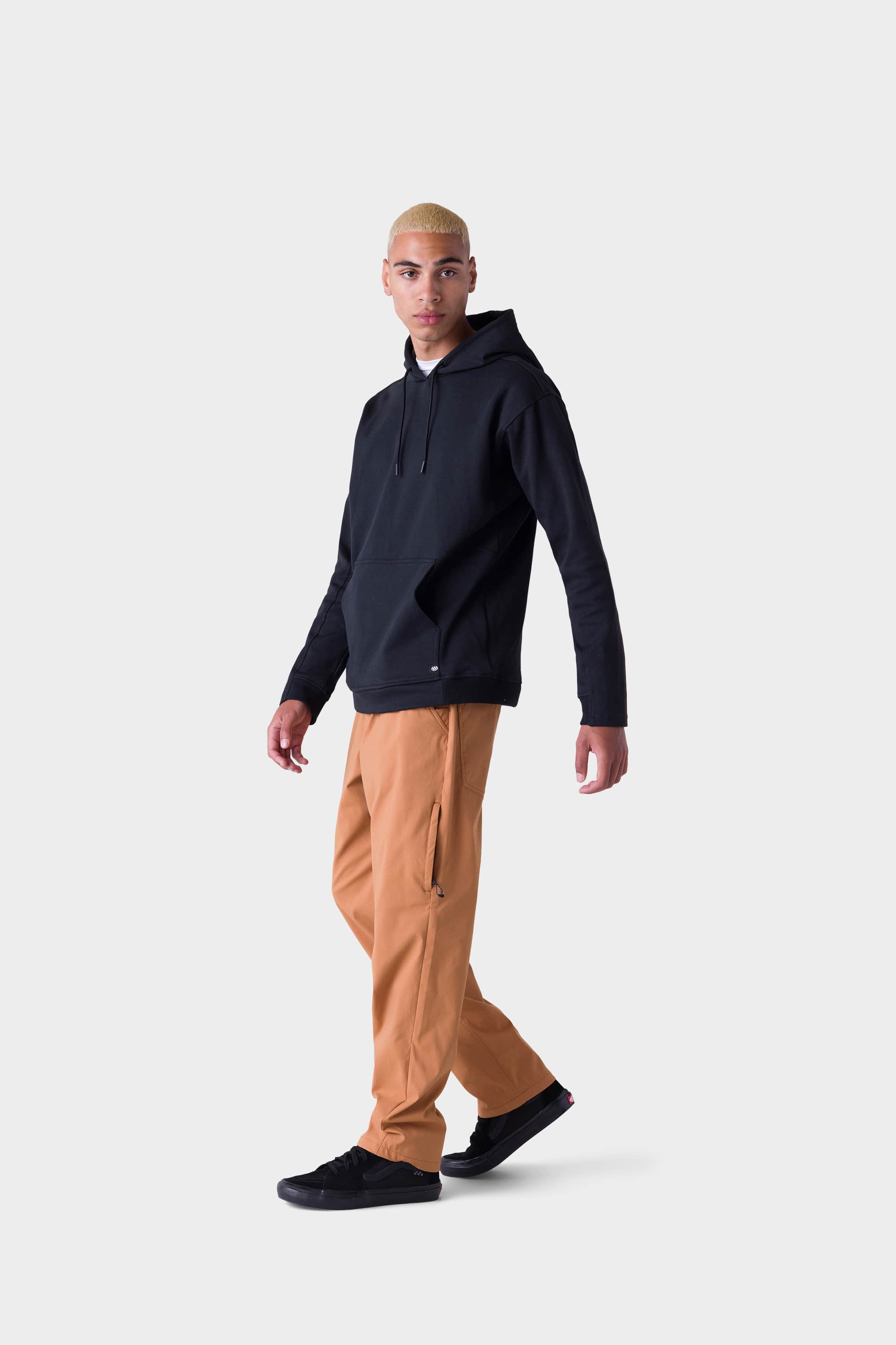 A young man with short blond hair wears a stylish insulated hoodie, 686 Men's Everywhere® Merino-Lined Pant - Relaxed Fit by 686, and black sneakers, walking against a plain light gray background.