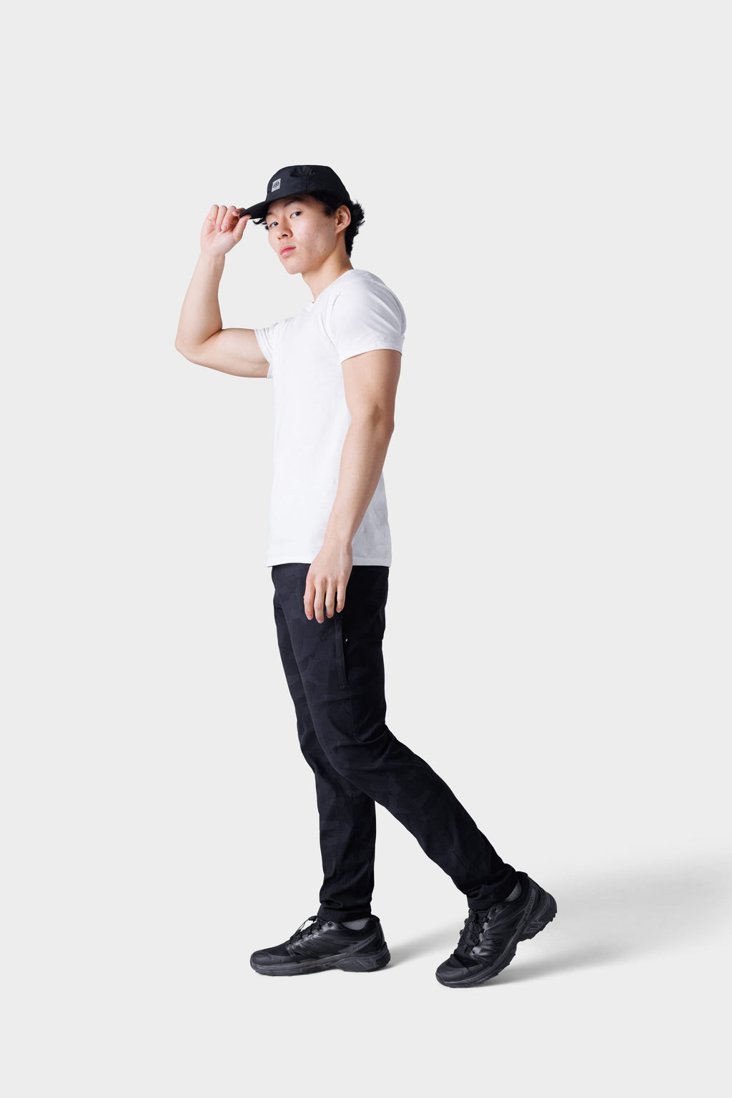 A man wears a white tee, black cap, black sneakers, and 686 Mens Everywhere Featherlight Chino Pant - Slim Fit, standing against a plain light gray background—an ideal look for summer adventures and warm weather style.