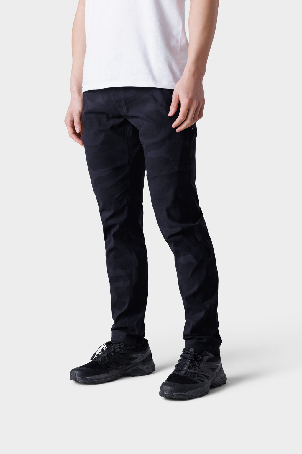 A person wearing a white t-shirt, 686 Mens Everywhere Featherlight Chino Pant - Slim Fit in black camo, and black sneakers stands against a light gray background. The image is cropped at the shoulders—perfect summer style inspiration.