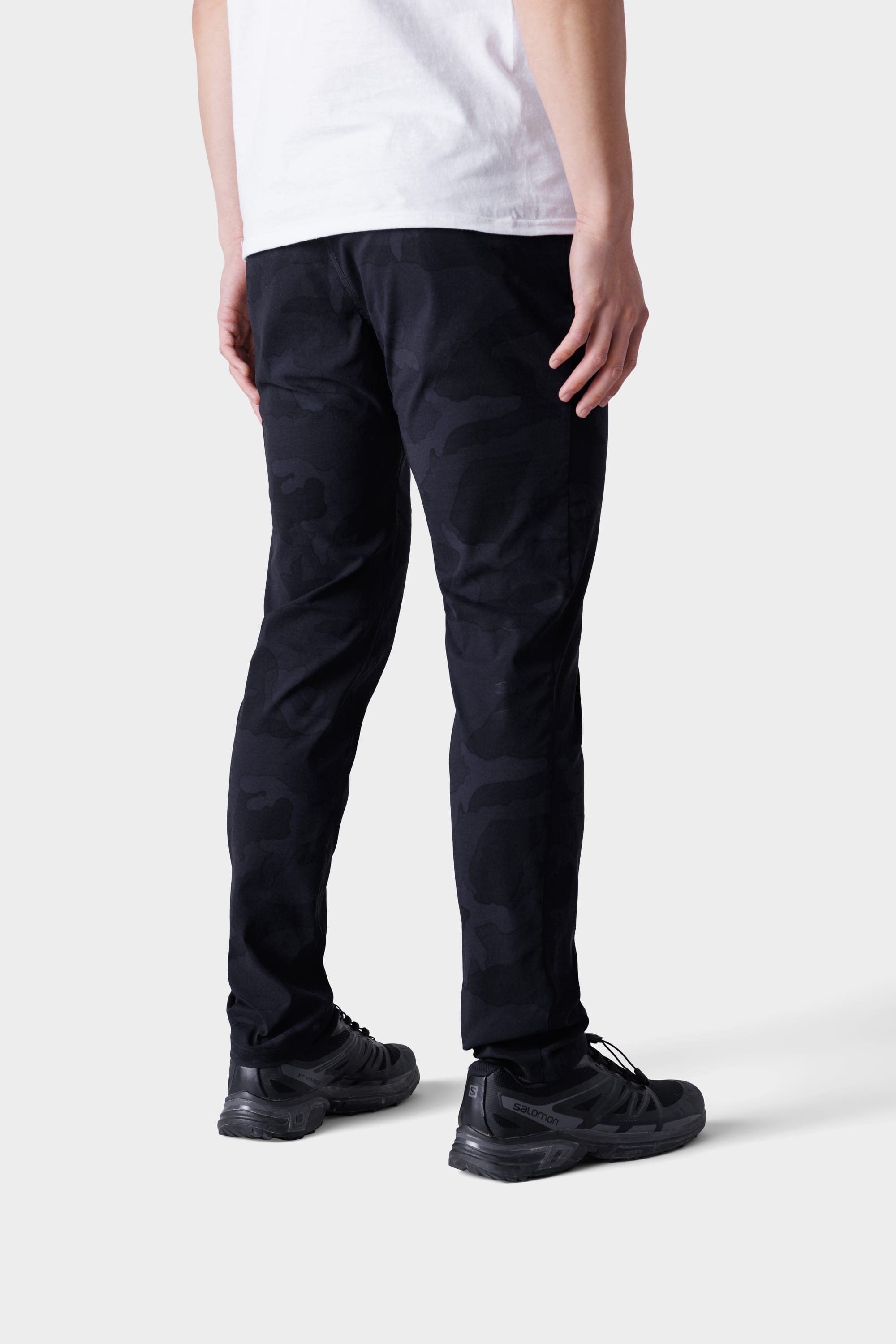 A person faces away, wearing a white t-shirt, 686 Mens Everywhere Featherlight Chino Pant - Slim Fit in black camo, and black sneakers against a plain light background—ideal for summer wardrobes or warm-weather outings.
