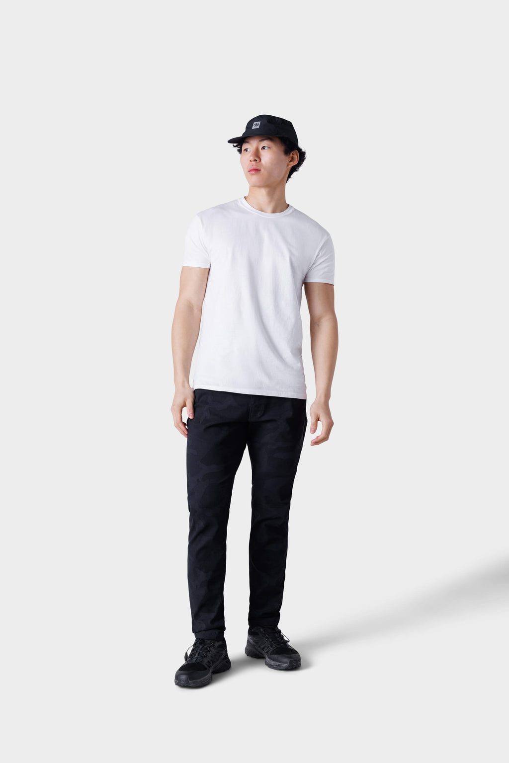 A person wearing a black bucket hat, plain white t-shirt, 686 Mens Everywhere Featherlight Chino Pant - Slim Fit in black camo, and black sneakers stands against a light gray background—perfect for warm weather adventures.