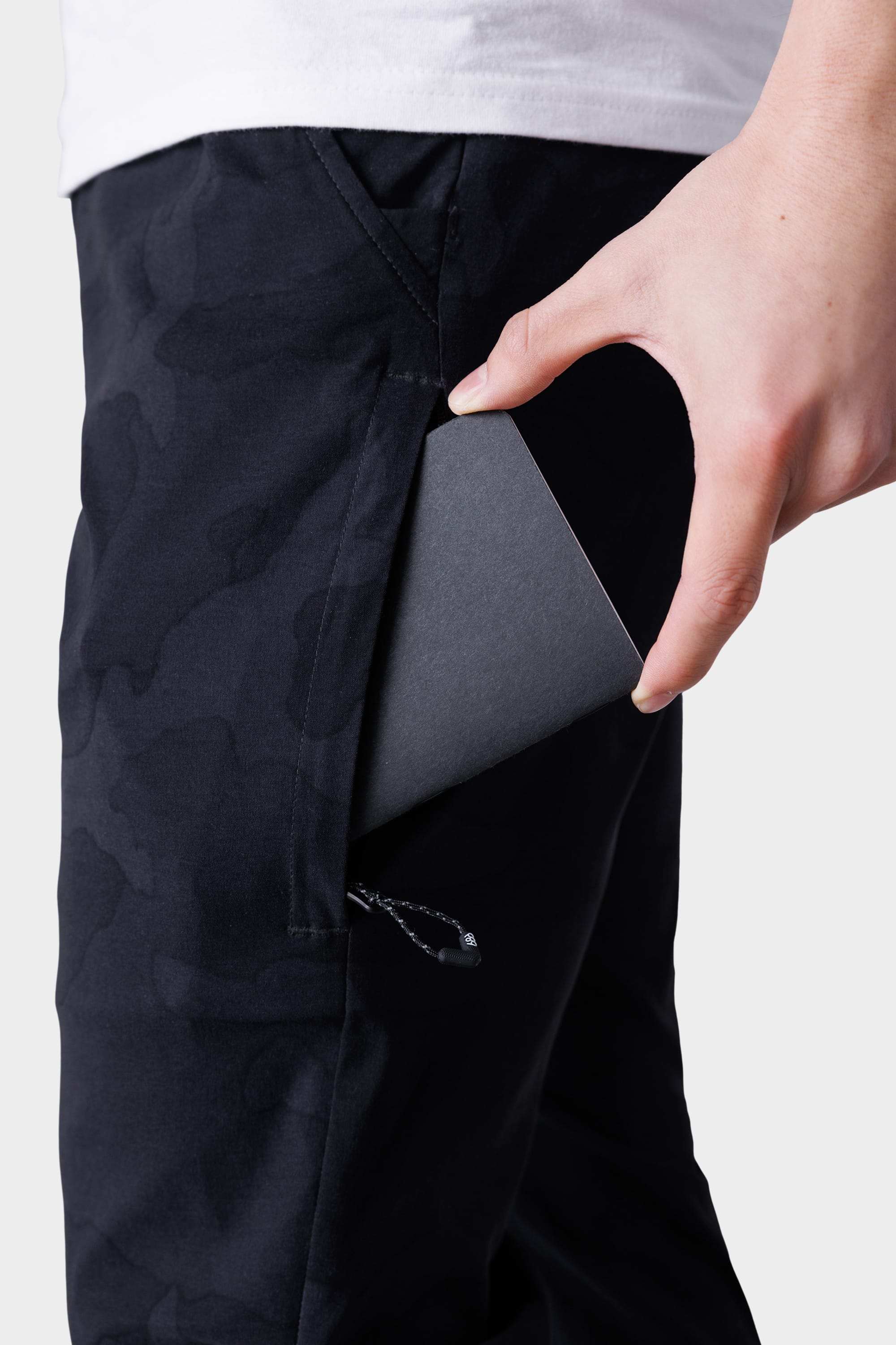 A person slides a slim, dark rectangle into the zippered side pocket of 686 Mens Everywhere Featherlight Chino Pant - Slim Fit by 686, featuring a black camo pattern ideal for warm-weather adventures.