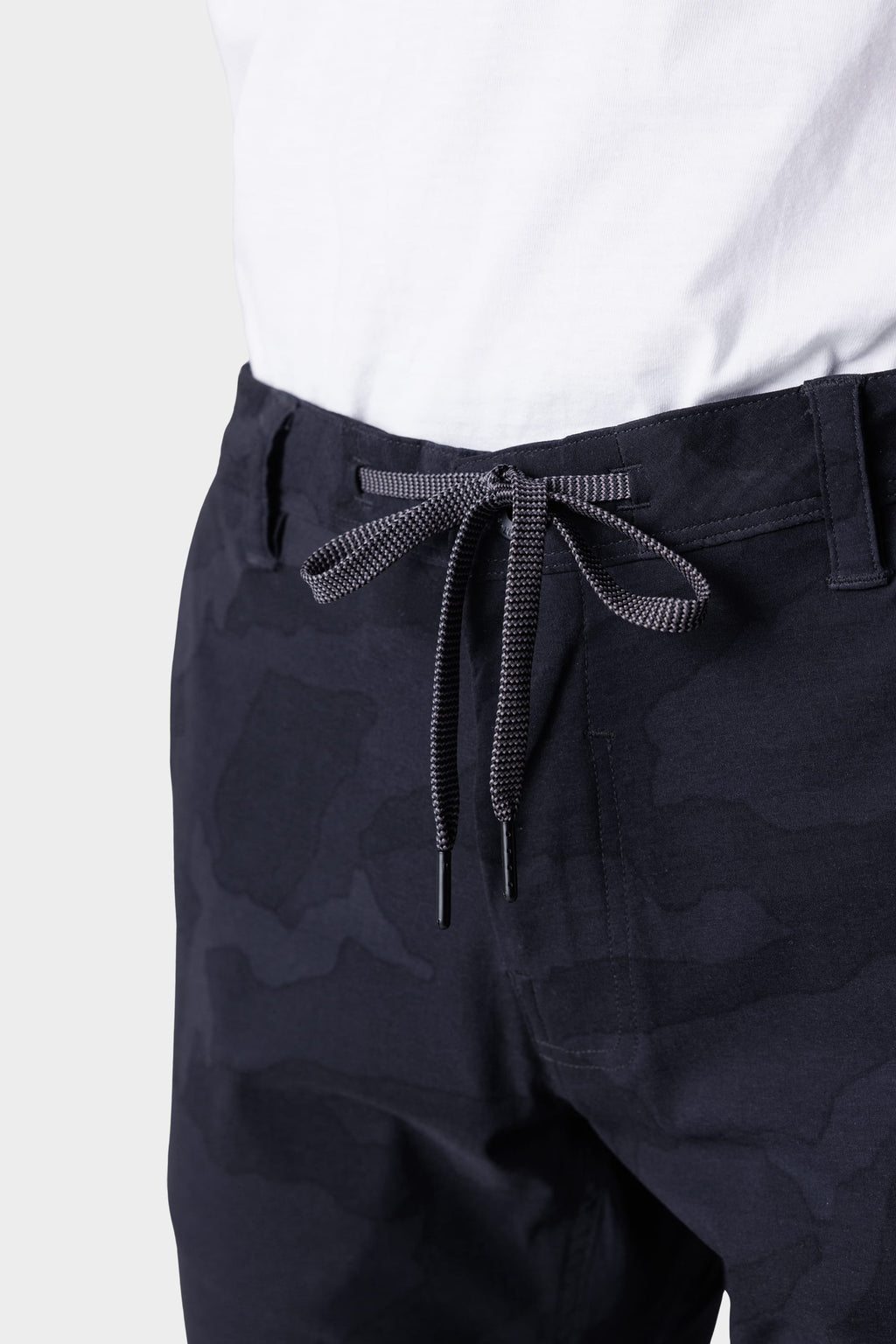A close-up of a person wearing a white shirt and 686 Mens Everywhere Featherlight Chino Pant - Slim Fit, featuring dark camouflage, lightweight fabric, and black-and-white drawstring ties—ideal for summer adventures or refreshing your warm-weather wardrobe.