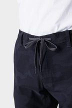 A close-up of a person wearing a white shirt and 686 Mens Everywhere Featherlight Chino Pant - Slim Fit, featuring dark camouflage, lightweight fabric, and black-and-white drawstring ties—ideal for summer adventures or refreshing your warm-weather wardrobe.