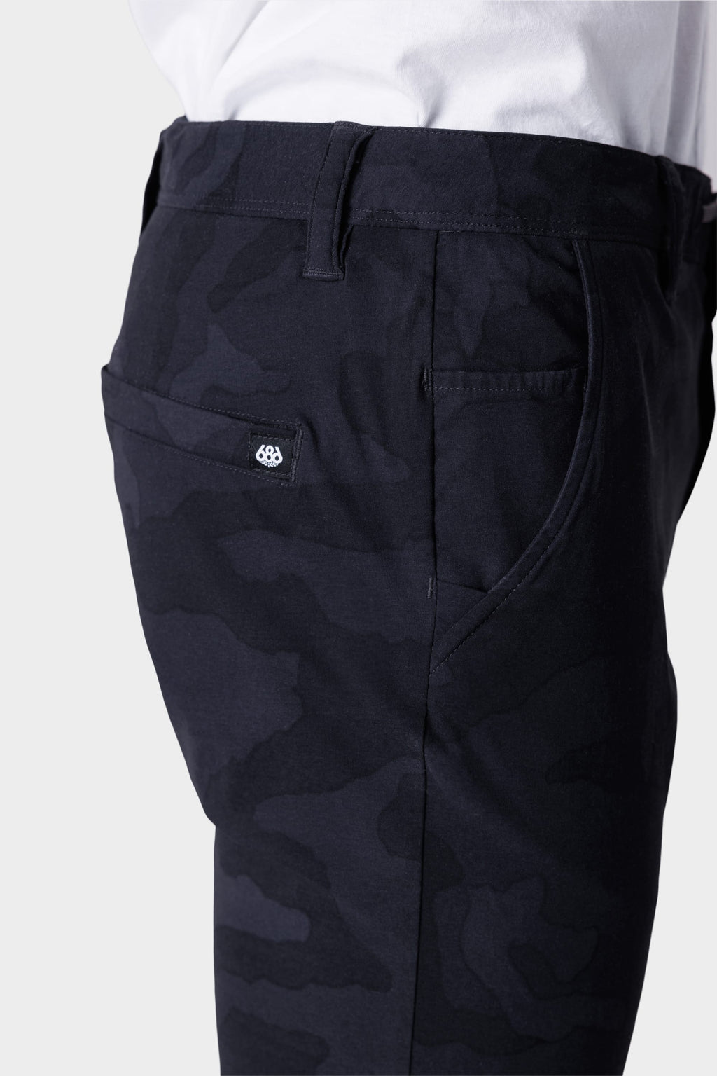 A close-up side view shows a person in 686 Mens Everywhere Featherlight Chino Pant - Slim Fit, featuring a camouflage pattern, back pocket with a fabric tag, and a white shirt—ideal for warm weather or updating your summer style.