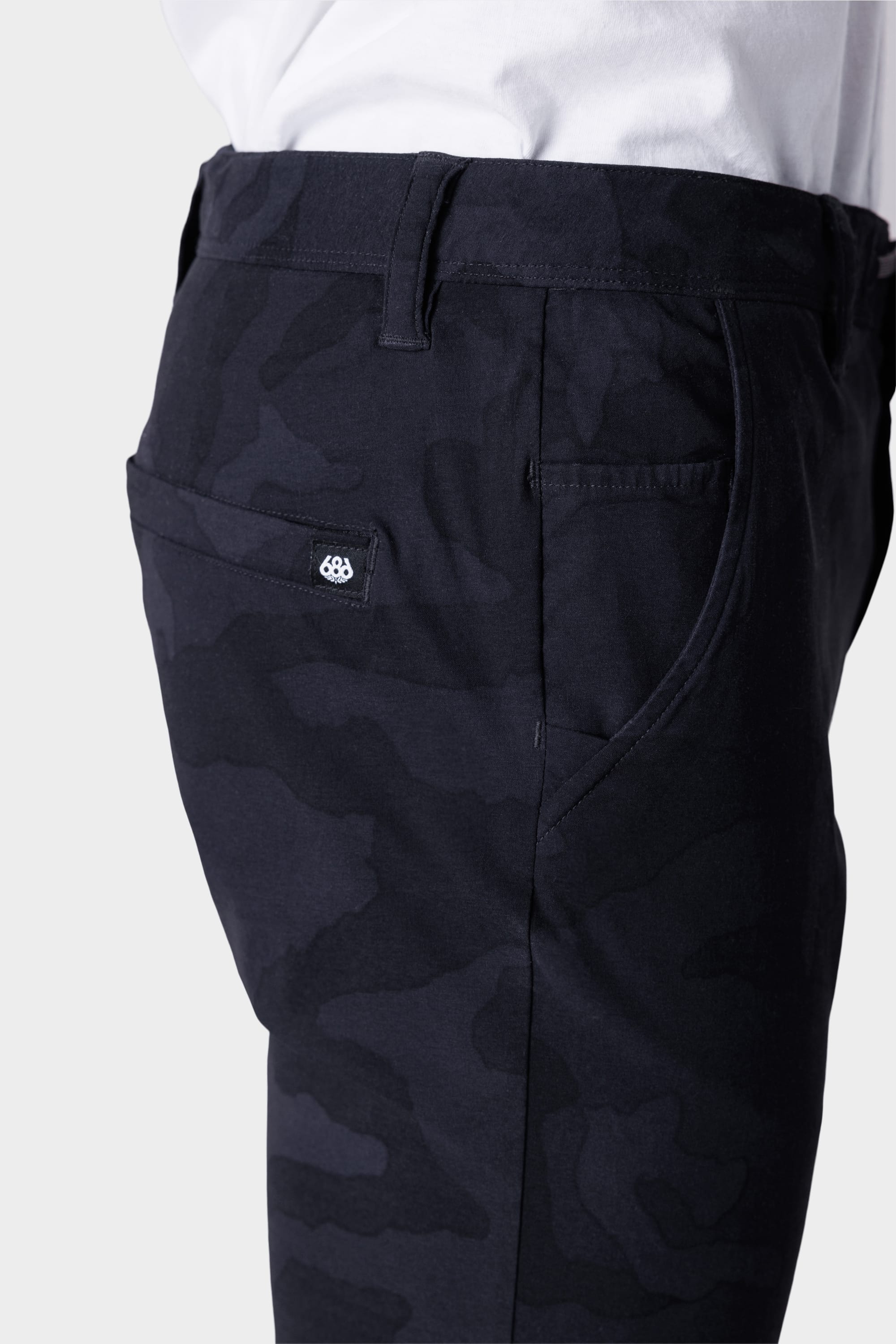 A close-up side view shows a person in 686 Mens Everywhere Featherlight Chino Pant - Slim Fit, featuring a camouflage pattern, back pocket with a fabric tag, and a white shirt—ideal for warm weather or updating your summer style.