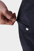 A hand pulls the zipper of a camo pocket on the 686 Mens Everywhere Featherlight Chino Pant - Slim Fit. A small white logo tag is near the pocket. The lightweight design is ideal for summer, shown against a plain light gray background.